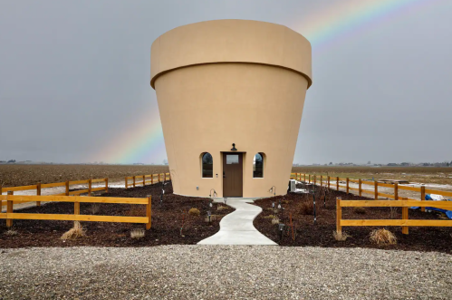 A unique, round building resembling a large pot, set in a field with a rainbow in the background and a gravel path leading to it.